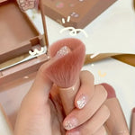 Makeup brush held by a hand with glittery nails, against a blurred makeup product background.